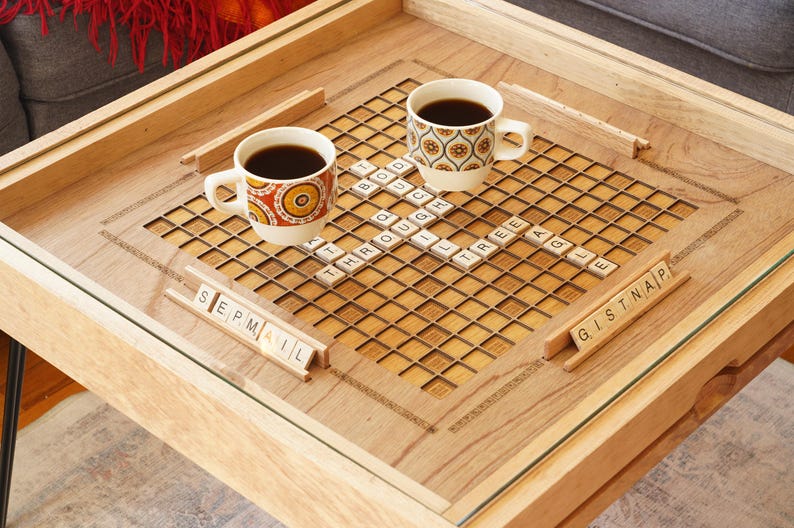 K&ouml;nnte beinhalten: Ein Holz-Couchtisch mit einer Glasplatte und einem eingebauten Scrabble-Brett. Zwei Tassen Kaffee stehen auf dem Brett, und es gibt Kacheln, die die W&ouml;rter "SEPMAIL" und "GISTNAIP" buchstabieren.