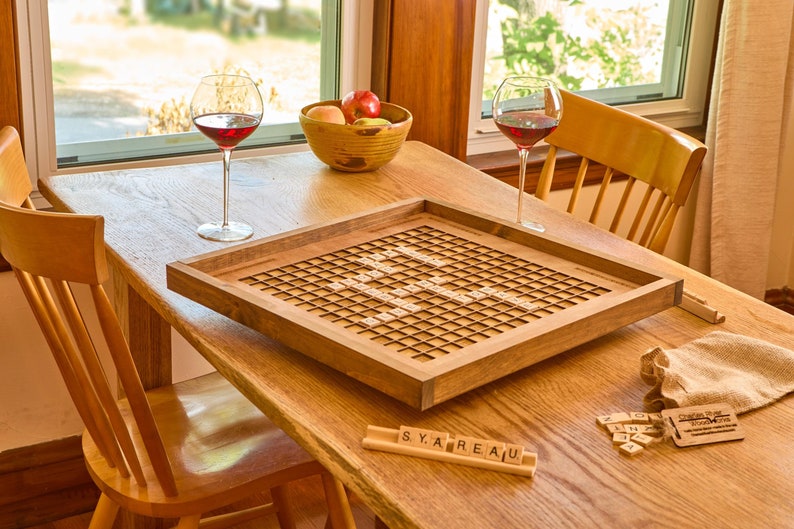 May include: A wooden game board with a grid pattern and a wooden frame. The board is set up for a game of Scrabble. There are two glasses of red wine on the table, along with a bowl of fruit. The game board is on a wooden table with two chairs.