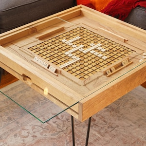 K&ouml;nnte beinhalten: Ein Holz-Couchtisch mit einer Glasplatte und einem eingebauten Scrabble-Spielbrett. Das Brett hat ein Gitter mit Quadraten und Kacheln mit Buchstaben darauf. Der Tisch hat schwarze Metallbeine.
