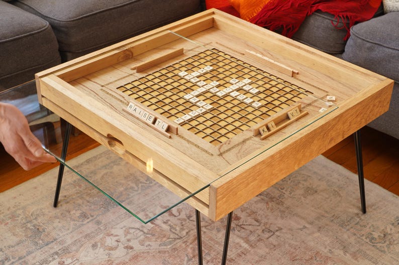 K&ouml;nnte beinhalten: Ein Holz-Couchtisch mit einer Glasplatte und einem eingebauten Scrabble-Spielbrett. Das Brett hat ein Gitter mit Quadraten und Kacheln mit Buchstaben darauf. Der Tisch hat schwarze Metallbeine.