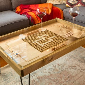 May include: A wooden coffee table with a clear glass top, featuring a Scrabble board design. The table has black hairpin legs and is surrounded by a gray sofa, pillows, and a rug. Two wine glasses with red wine are on the table.