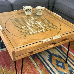 May include: Wooden coffee table with a built-in game board and glass top. The table has black hairpin legs and includes two decorative mugs. The words "VER" and "YORE" are visible on the table.