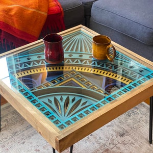 May include: A wooden coffee table with a glass top featuring a blue and yellow geometric design. The table has four black hairpin legs and is decorated with a red ceramic pitcher and a yellow ceramic mug.