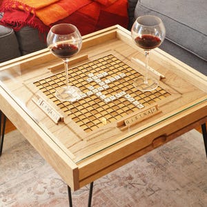 K&ouml;nnte beinhalten: Ein Holz-Couchtisch mit einer Glasplatte und einem eingebauten Scrabble-Spielfeld. Der Tisch hat schwarze Haarnadelbeine und zwei Weingl&auml;ser stehen auf dem Tisch.