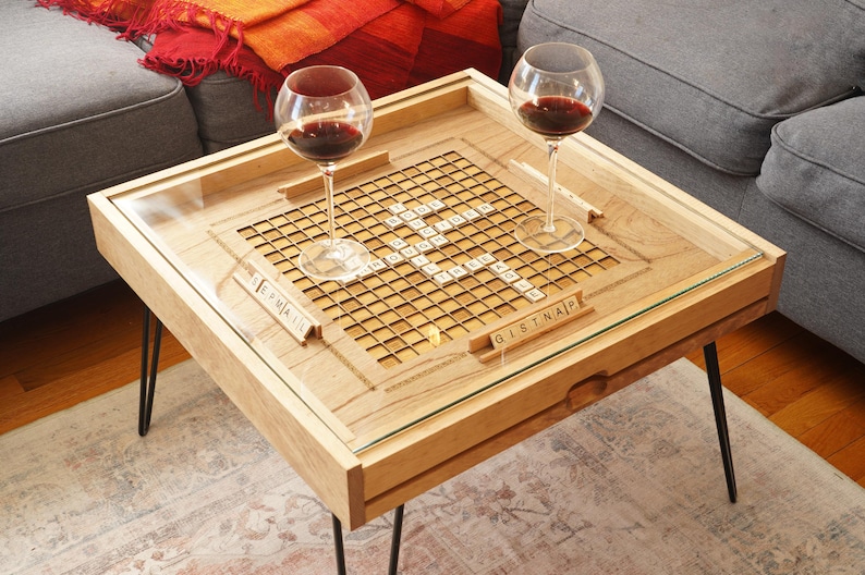K&ouml;nnte beinhalten: Ein Holz-Couchtisch mit einer Glasplatte und einem eingebauten Scrabble-Spielfeld. Der Tisch hat schwarze Haarnadelbeine und zwei Weingl&auml;ser stehen auf dem Tisch.