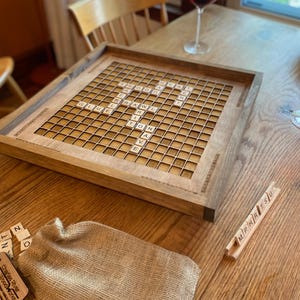 May include: A wooden game board with a Scrabble game set up on it. The board is made of walnut wood and has a natural finish. The game pieces are arranged in a grid pattern on the board. There are two glasses of red wine on the table next to the board.
