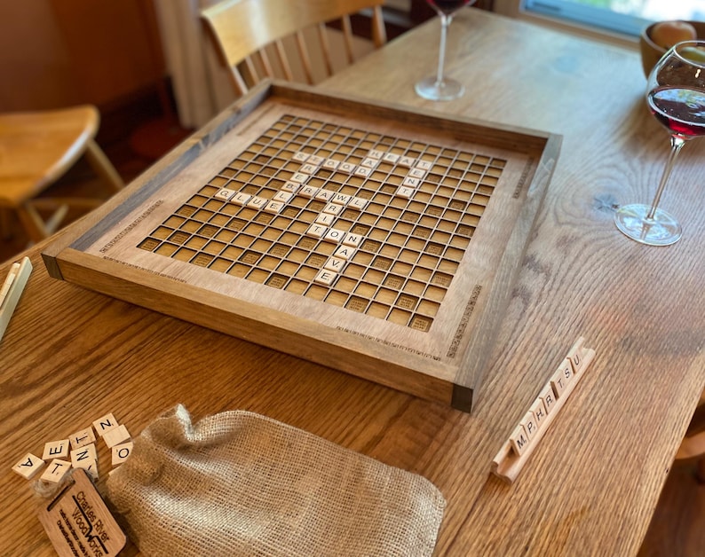May include: A wooden game board with a Scrabble game set up on it. The board is made of walnut wood and has a natural finish. The game pieces are arranged in a grid pattern on the board. There are two glasses of red wine on the table next to the board.