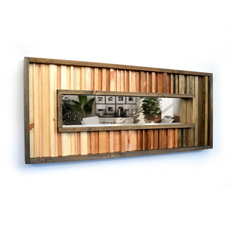 Puede incluir: Un espejo rectangular de madera con un dise&ntilde;o &uacute;nico que presenta una variedad de tonos y texturas de madera. El espejo tiene una abertura rectangular estrecha en el centro, que revela un reflejo de una habitaci&oacute;n con plantas y muebles.