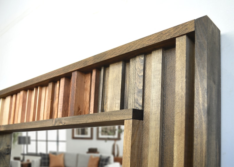 Puede incluir: Un espejo rectangular con un marco &uacute;nico hecho de diferentes tonos de madera. El marco est&aacute; hecho de finas tiras de madera dispuestas en un patr&oacute;n vertical, creando un efecto de capas. El espejo est&aacute; rodeado por un borde grueso de madera marr&oacute;n oscuro.