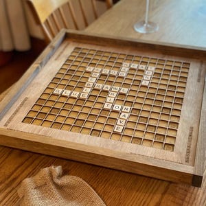 May include: A wooden Scrabble board with a light brown finish. The board has a grid of 15x15 squares with a dark brown border. The board is set up for a game of Scrabble, with tiles placed on the board to form words. The tiles are made of wood and have white letters on them.