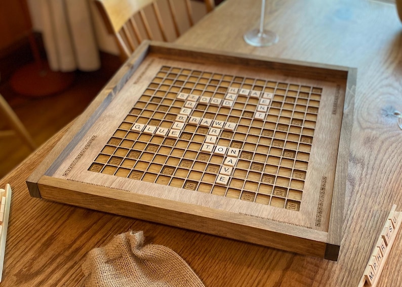 May include: A wooden Scrabble board with a light brown finish. The board has a grid of 15x15 squares with a dark brown border. The board is set up for a game of Scrabble, with tiles placed on the board to form words. The tiles are made of wood and have white letters on them.