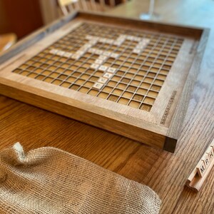 May include: A wooden Scrabble game board with a light brown finish. The board has a grid of 15x15 squares, with a center star and letter tiles in the squares. The board is on a wooden table with a burlap bag and a rack of letter tiles.