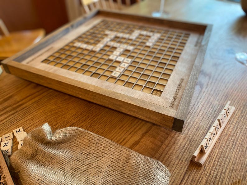 May include: A wooden Scrabble game board with a light brown finish. The board has a grid of 15x15 squares, with a center star and letter tiles in the squares. The board is on a wooden table with a burlap bag and a rack of letter tiles.