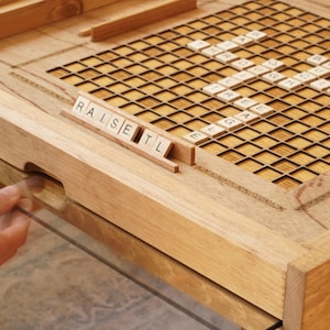 K&ouml;nnte beinhalten: Ein h&ouml;lzernes Spielbrett mit einem Gitter aus Quadraten und Buchstabenpl&auml;ttchen, die die W&ouml;rter "raise", "set" und "tl" buchstabieren.