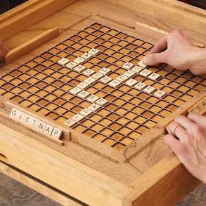 K&ouml;nnte beinhalten: Ein h&ouml;lzernes Spielbrett mit einem Gitter aus Quadraten, gef&uuml;llt mit Buchstabenkacheln, die W&ouml;rter wie "Glider", "Quig" und "Tree" buchstabieren.