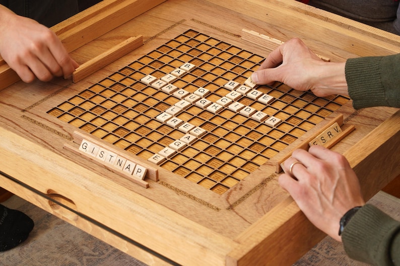 K&ouml;nnte beinhalten: Ein h&ouml;lzernes Spielbrett mit einem Gitter aus Quadraten, gef&uuml;llt mit Buchstabenkacheln, die W&ouml;rter wie "Glider", "Quig" und "Tree" buchstabieren.