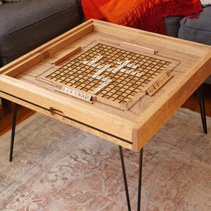 K&ouml;nnte beinhalten: Ein h&ouml;lzerner Spieltisch mit schwarzen Haarnadelbeinen. Die Tischplatte hat ein eingebautes Scrabble-Brett mit Kacheln, die "GISTNAP" und "KILLERBEE" buchstabieren.