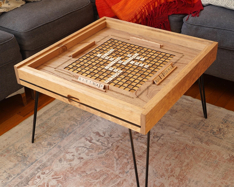 K&ouml;nnte beinhalten: Ein h&ouml;lzerner Spieltisch mit schwarzen Haarnadelbeinen. Die Tischplatte hat ein eingebautes Scrabble-Brett mit Kacheln, die "GISTNAP" und "KILLERBEE" buchstabieren.