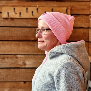 May include: A light pink beanie with a small heart detail is worn by a person wearing glasses and a gray fleece jacket with a pink zipper. The background is a wooden wall with hooks.