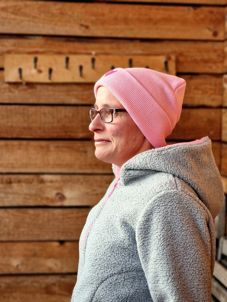 May include: A light pink beanie with a small heart detail is worn by a person wearing glasses and a gray fleece jacket with a pink zipper. The background is a wooden wall with hooks.