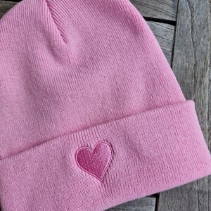 May include: A light pink beanie with a folded brim. A darker pink heart is embroidered on the front of the hat. The hat is made of a soft, knitted material.