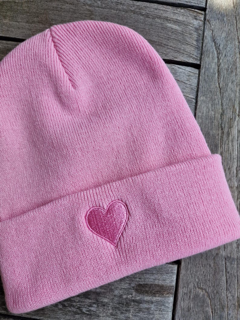 May include: A light pink beanie with a folded brim. A darker pink heart is embroidered on the front of the hat. The hat is made of a soft, knitted material.
