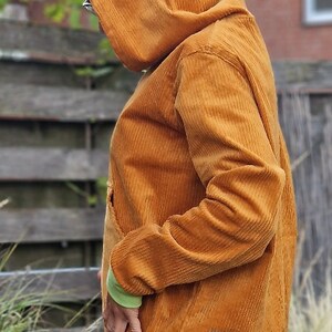 May include: A mustard yellow corduroy hooded sweatshirt featuring long sleeves with light green cuffs, a front kangaroo pocket, and a solid mustard yellow hem. A small rectangular teal fabric patch with white text is stitched near the bottom hem.
