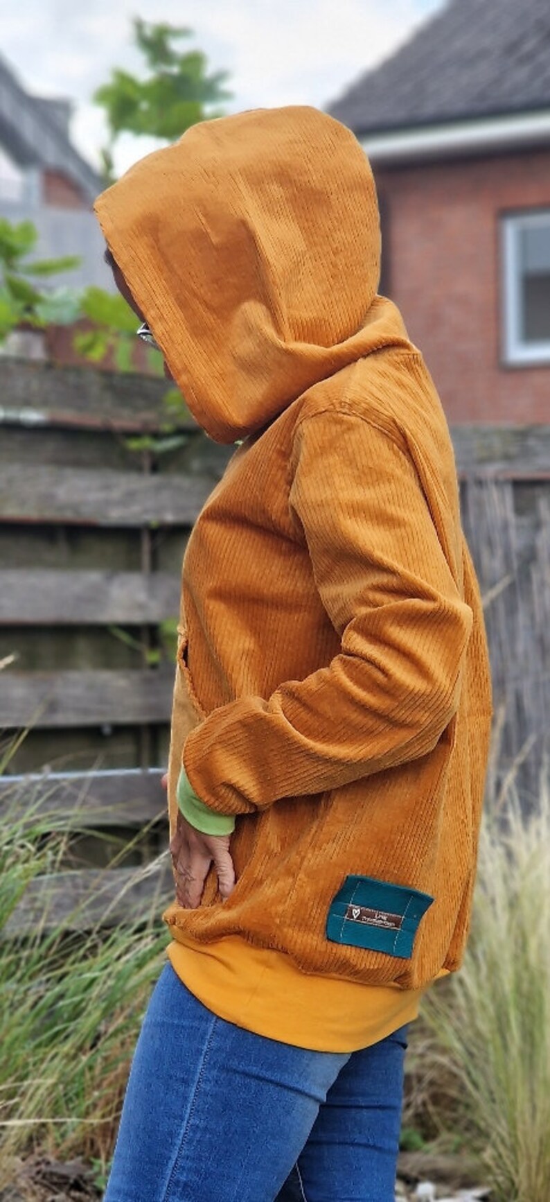 May include: A mustard yellow corduroy hooded sweatshirt featuring long sleeves with light green cuffs, a front kangaroo pocket, and a solid mustard yellow hem. A small rectangular teal fabric patch with white text is stitched near the bottom hem.