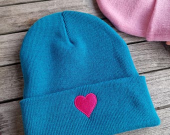 May include: A teal beanie with a pink heart embroidered on the front. The folded brim and the heart are a vibrant pink. Another pink beanie is partially visible in the background. The beanies are made of a soft knit material.