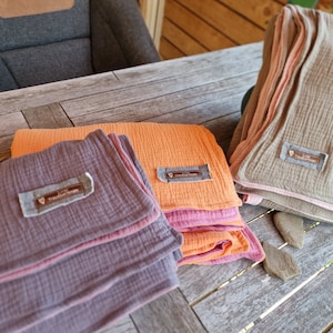 Pu&ograve; includere: Tre coperte di mussola piegate in diversi colori: grigio, arancione e rosa. Ogni coperta ha una piccola etichetta con il testo "Love, Treasured Moments".