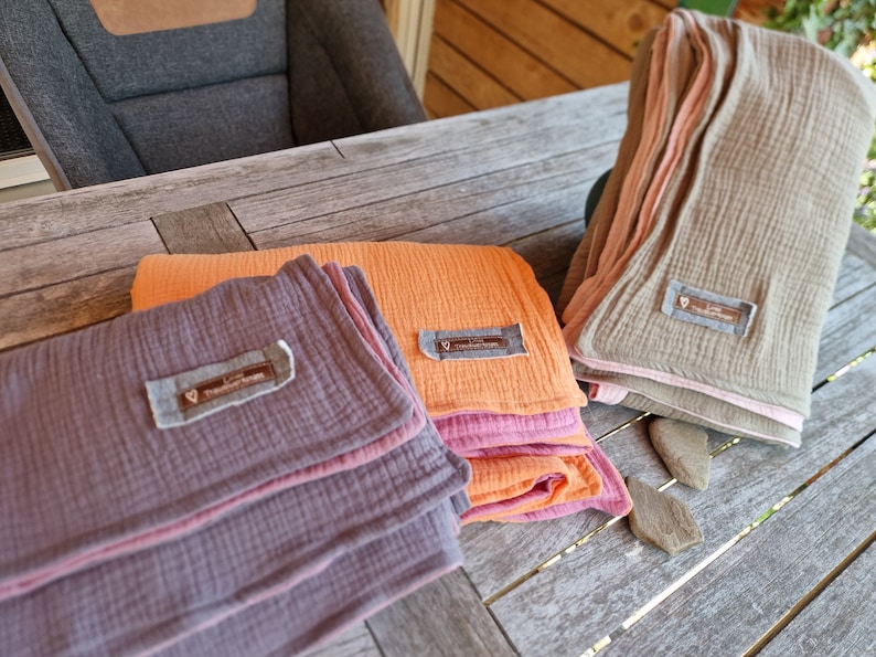 Pu&ograve; includere: Tre coperte di mussola piegate in diversi colori: grigio, arancione e rosa. Ogni coperta ha una piccola etichetta con il testo "Love, Treasured Moments".