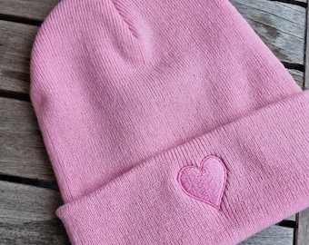 May include: A pink beanie hat with a folded brim. The hat features a stitched heart emblem on the front. The hat is made of a soft, knitted material. The beanie is a casual accessory.