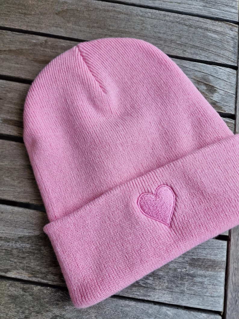 May include: A pink beanie hat with a folded brim. The hat features a stitched heart emblem on the front. The hat is made of a soft, knitted material. The beanie is a casual accessory.
