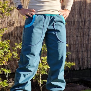 Könnte beinhalten: Eine Hose in Tealblau mit türkisfarbenem Besatz an den Taschen und am Bund. Die Hose ist an den Knöcheln gerafft und die Person, die sie trägt, steht auf einem Rasen.