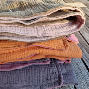 Pu&ograve; includere: Un mucchio di quattro coperte in mussola di cotone di diversi colori: beige, rosa, arancio e viola. Le coperte sono piegate e hanno un bordo cucito decorativo.