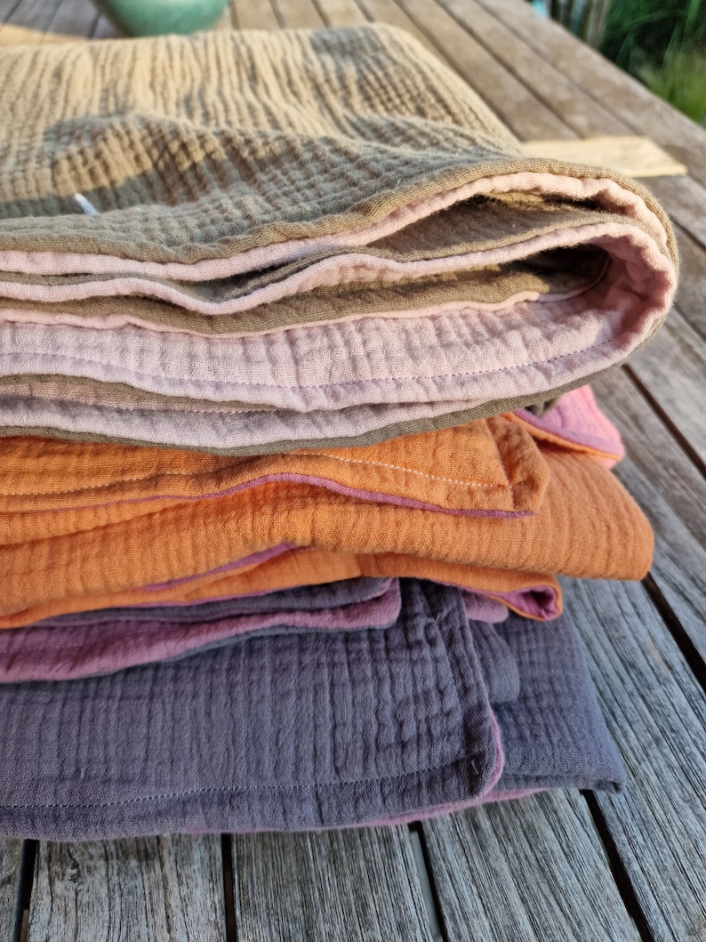 Pu&ograve; includere: Un mucchio di quattro coperte in mussola di cotone di diversi colori: beige, rosa, arancio e viola. Le coperte sono piegate e hanno un bordo cucito decorativo.