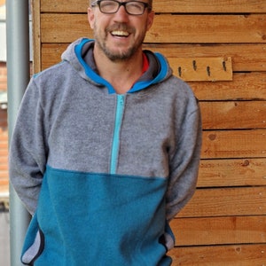 May include: A man wearing a gray and blue hooded sweatshirt with a zipper. He is smiling and standing in front of a wooden wall.