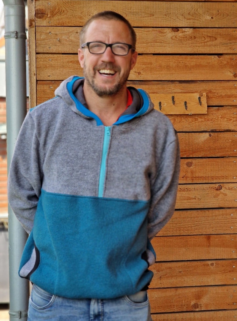 May include: A man wearing a gray and blue hooded sweatshirt with a zipper. He is smiling and standing in front of a wooden wall.