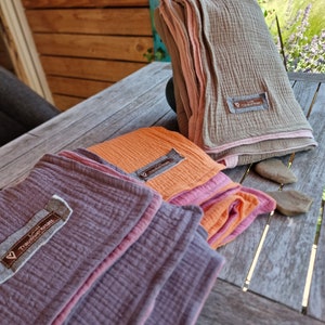 Pu&ograve; includere: Tre coperte di mussola piegate in diversi colori: viola, arancione e beige. Ogni coperta ha un bordo rosa e una piccola etichetta con il testo "Lieb Tr&auml;umedecke" su di essa.