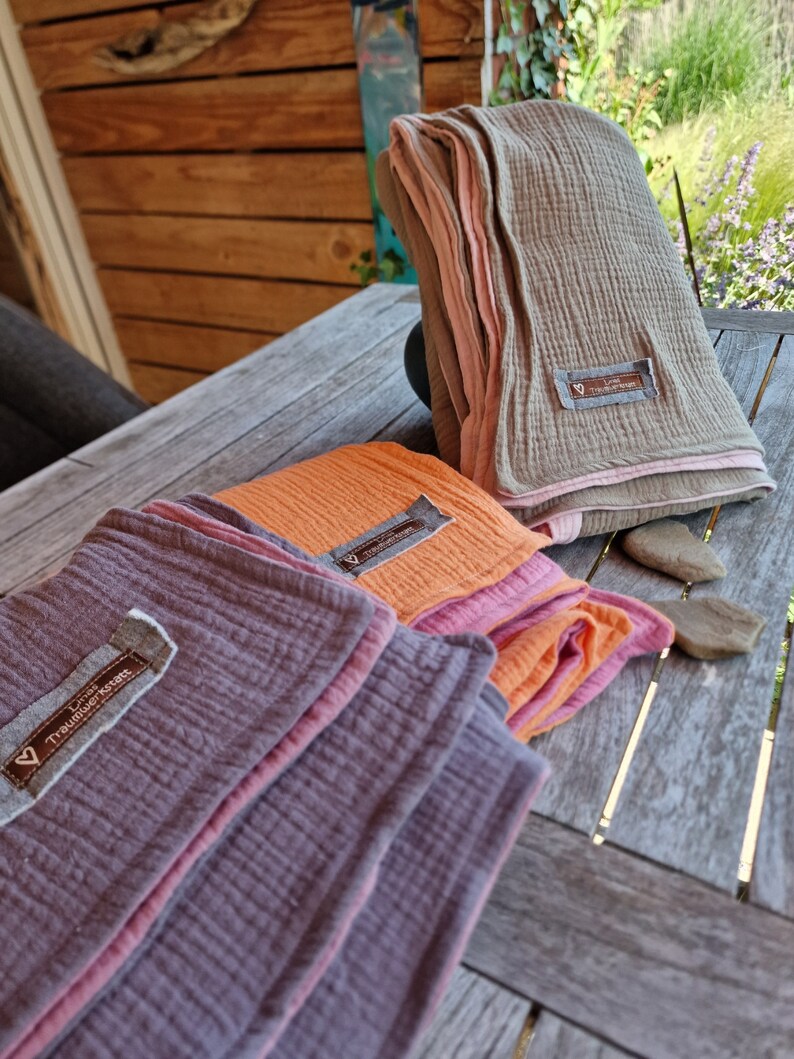 Pu&ograve; includere: Tre coperte di mussola piegate in diversi colori: viola, arancione e beige. Ogni coperta ha un bordo rosa e una piccola etichetta con il testo "Lieb Tr&auml;umedecke" su di essa.