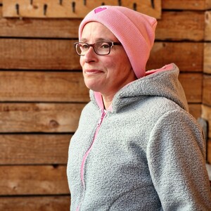 May include: A woman wearing a gray fleece hooded jacket with a pink beanie featuring a pink heart. The beanie has a ribbed texture.