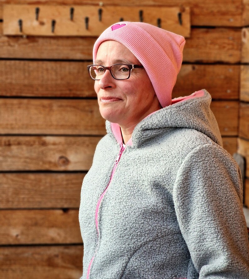 May include: A woman wearing a gray fleece hooded jacket with a pink beanie featuring a pink heart. The beanie has a ribbed texture.