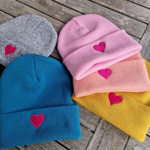 May include: A collection of colorful knit beanies. The beanies are in shades of gray, teal, pink, peach, and yellow. Each beanie features a bright pink heart embroidered on the front. The hats are displayed on a wooden surface.