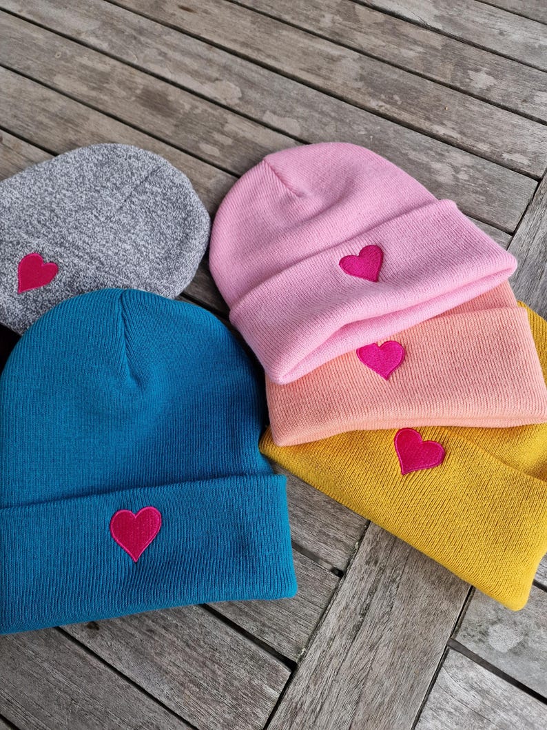 May include: A collection of colorful knit beanies. The beanies are in shades of gray, teal, pink, peach, and yellow. Each beanie features a bright pink heart embroidered on the front. The hats are displayed on a wooden surface.
