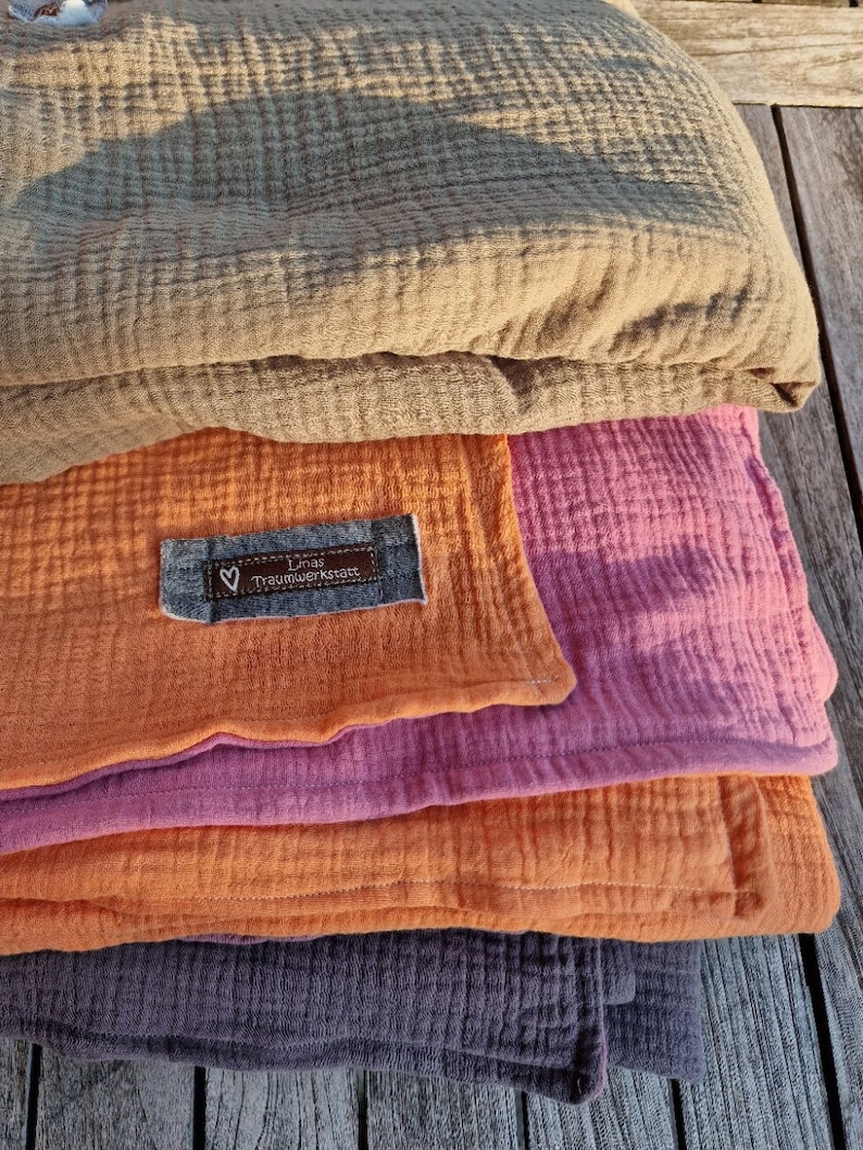 Pu&ograve; includere: Un mucchio di quattro coperte di mussola in diverse tonalit&agrave; di marrone, rosa, arancione e viola. La coperta superiore ha un'etichetta piccola che recita "Lina's Traumwerkstatt".