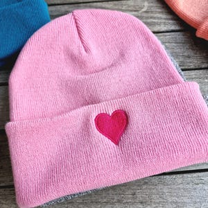 May include: A pink beanie with a gray brim and a red heart embroidered on the front.