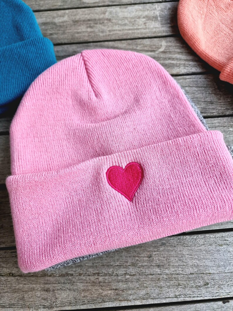 May include: A pink beanie with a gray brim and a red heart embroidered on the front.