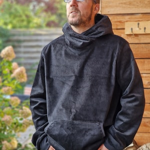 Könnte beinhalten: Ein schwarzer Cord-Hoodie mit hohem Kragen und einer Kängurutasche. Der Hoodie wird von einer Person getragen, die eine blaue Jeans trägt.