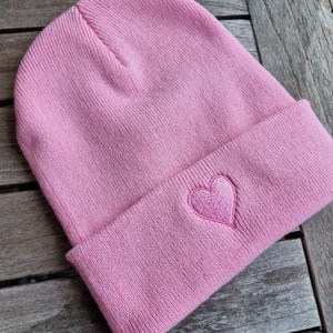 May include: A light pink beanie with a folded brim. A heart is embroidered on the folded brim. The beanie is made of a soft knit material.