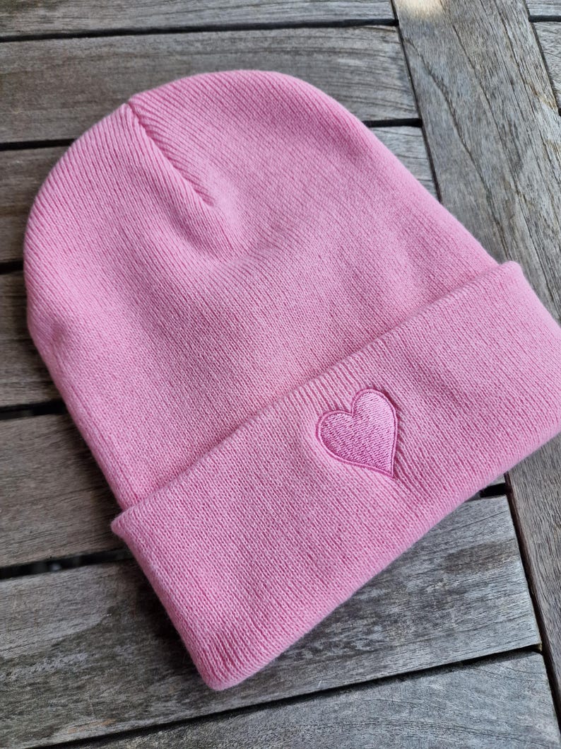 May include: A light pink beanie with a folded brim. A heart is embroidered on the folded brim. The beanie is made of a soft knit material.